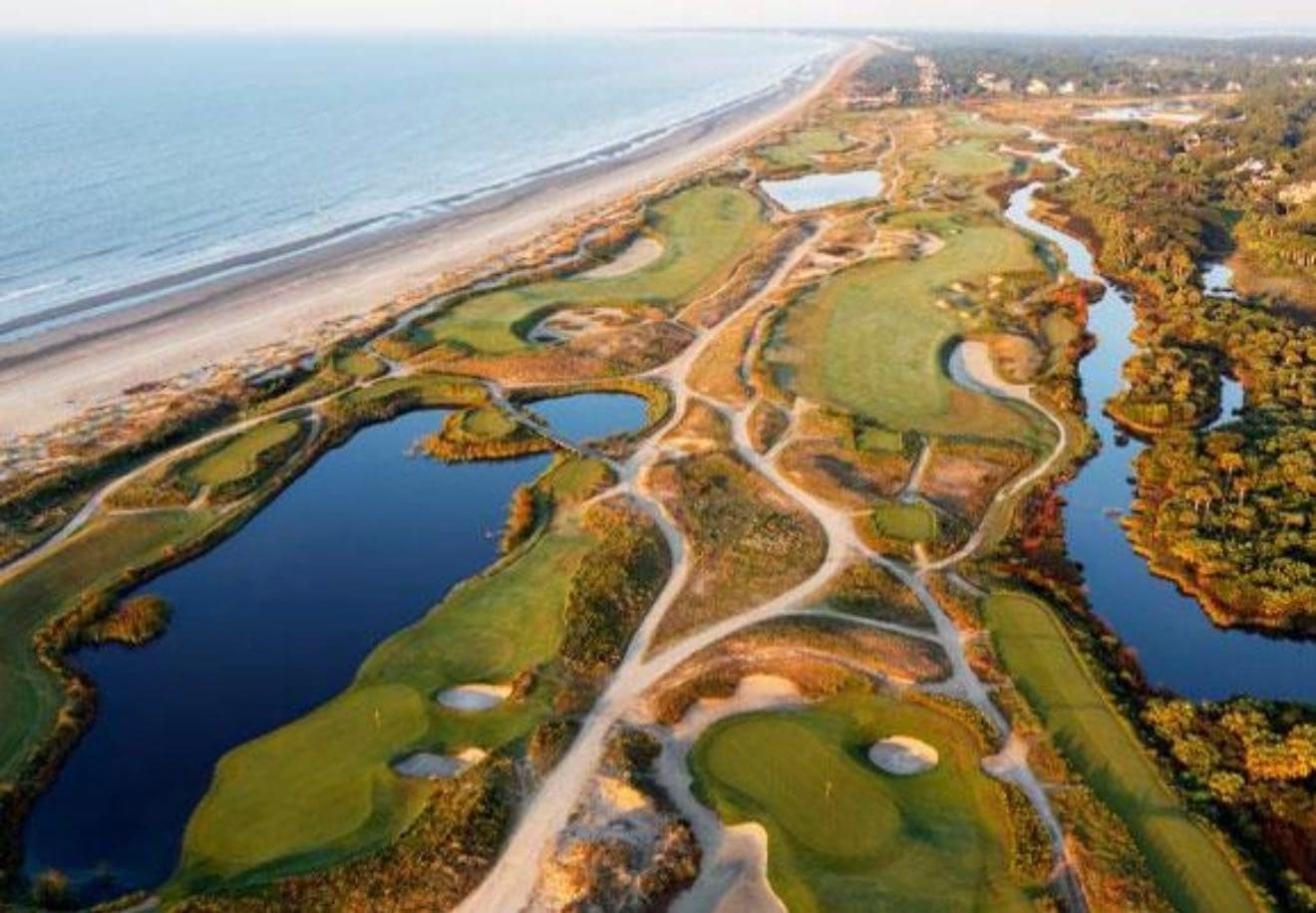 Aerial view of an oceanfront golf course winding through dunes, lagoons and maritime forest beside a long, wide beach