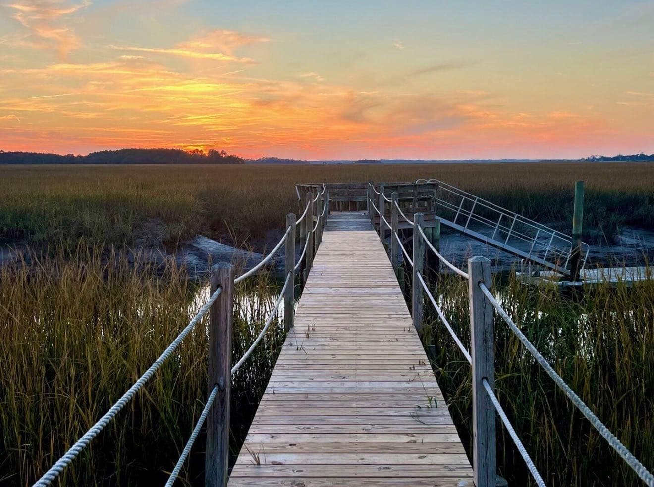 Wooden dock with rope rails running out over spartina marsh and a tidal creek at sunset